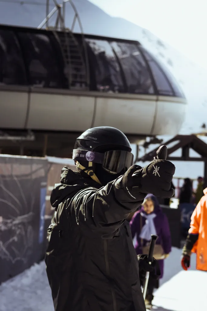 Person in snow gear and helmet pointing toward the camera beside a cable car